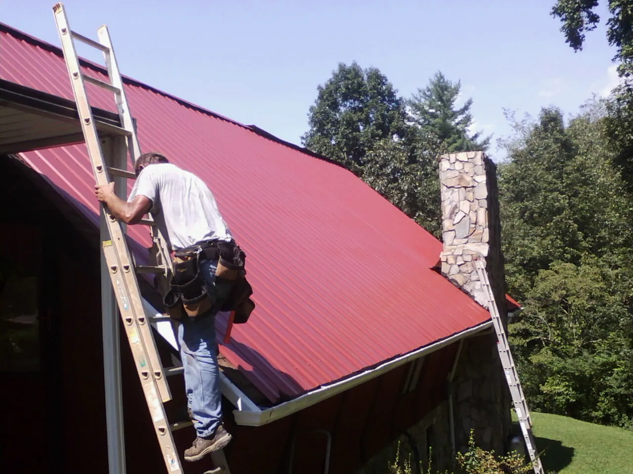 Roofer on a ladder beside a red metal roof and stone chimney — Soffit & Fascia Repair by Roofing Brothers in Geneseo
